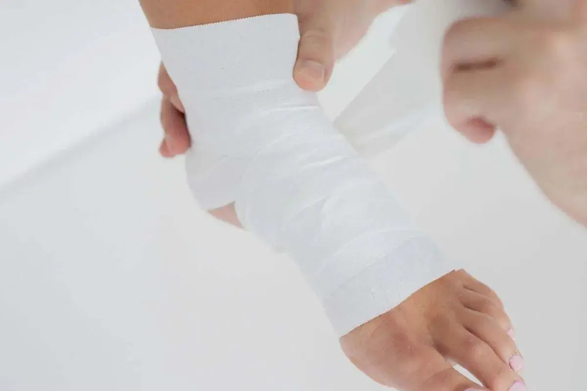 Dłonie owijają stopę białą taśmą do tapingu, zapewniając wsparcie i stabilizację.