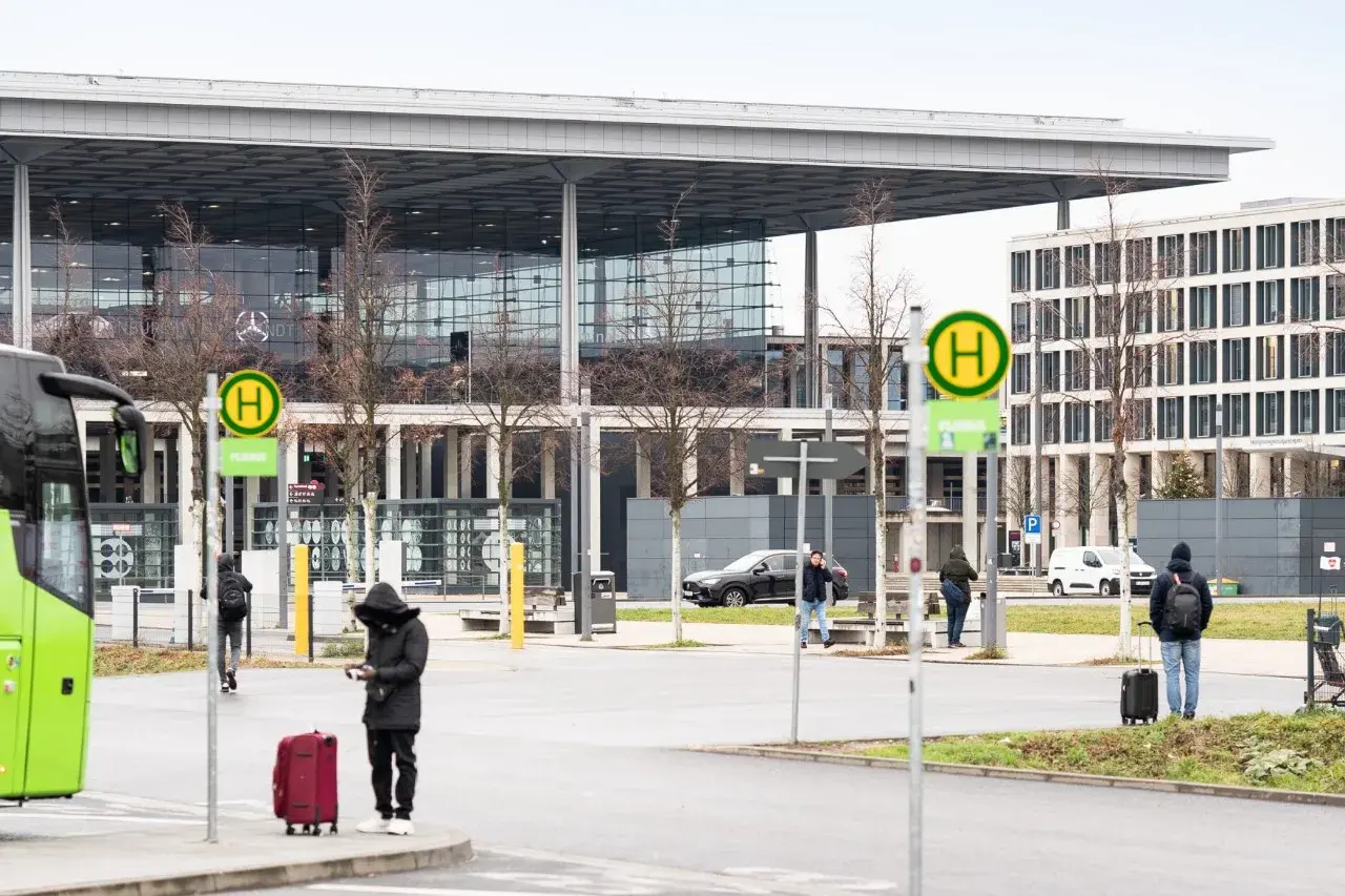Przystanek autobusowy z zielonym autobusem, ludźmi z bagażami i nowoczesnym budynkiem w tle.