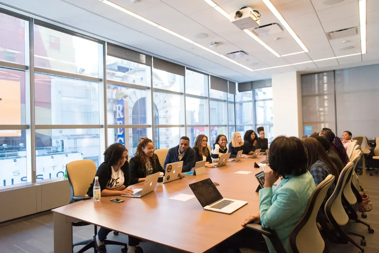 A diverse group of professionals engage in government leadership training, collaborating around a conference table with laptops and tablets.