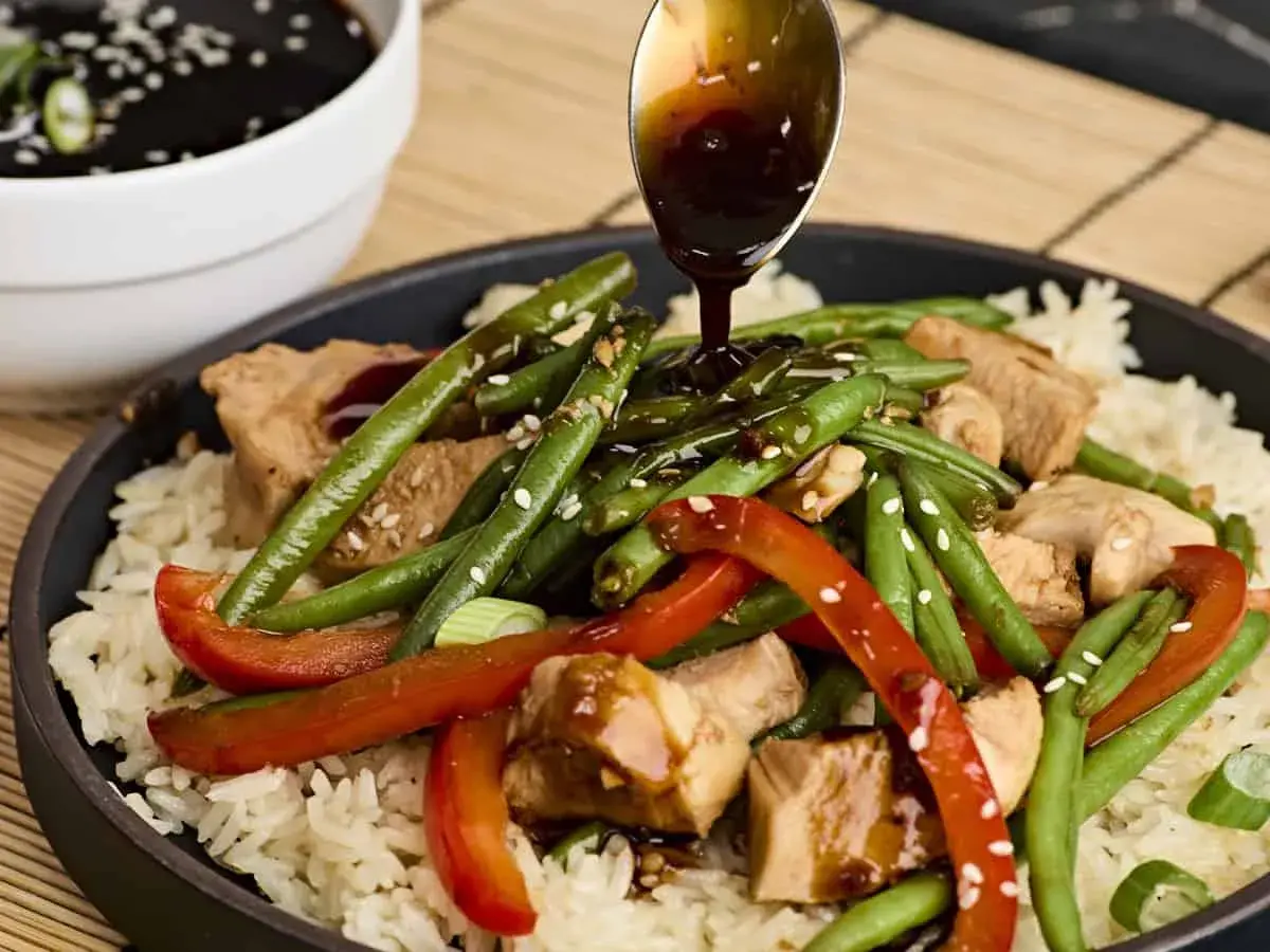 A spoon drizzles teriyaki sauce over a dish of rice, chicken, green beans, and red peppers, garnished with sesame seeds.