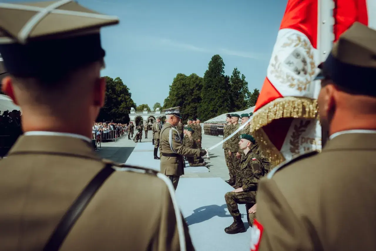 Podczas uroczystości wojskowej, oficer wręcza szablę klęczącemu podporucznikowi. W tle stoją żołnierze i widzowie.