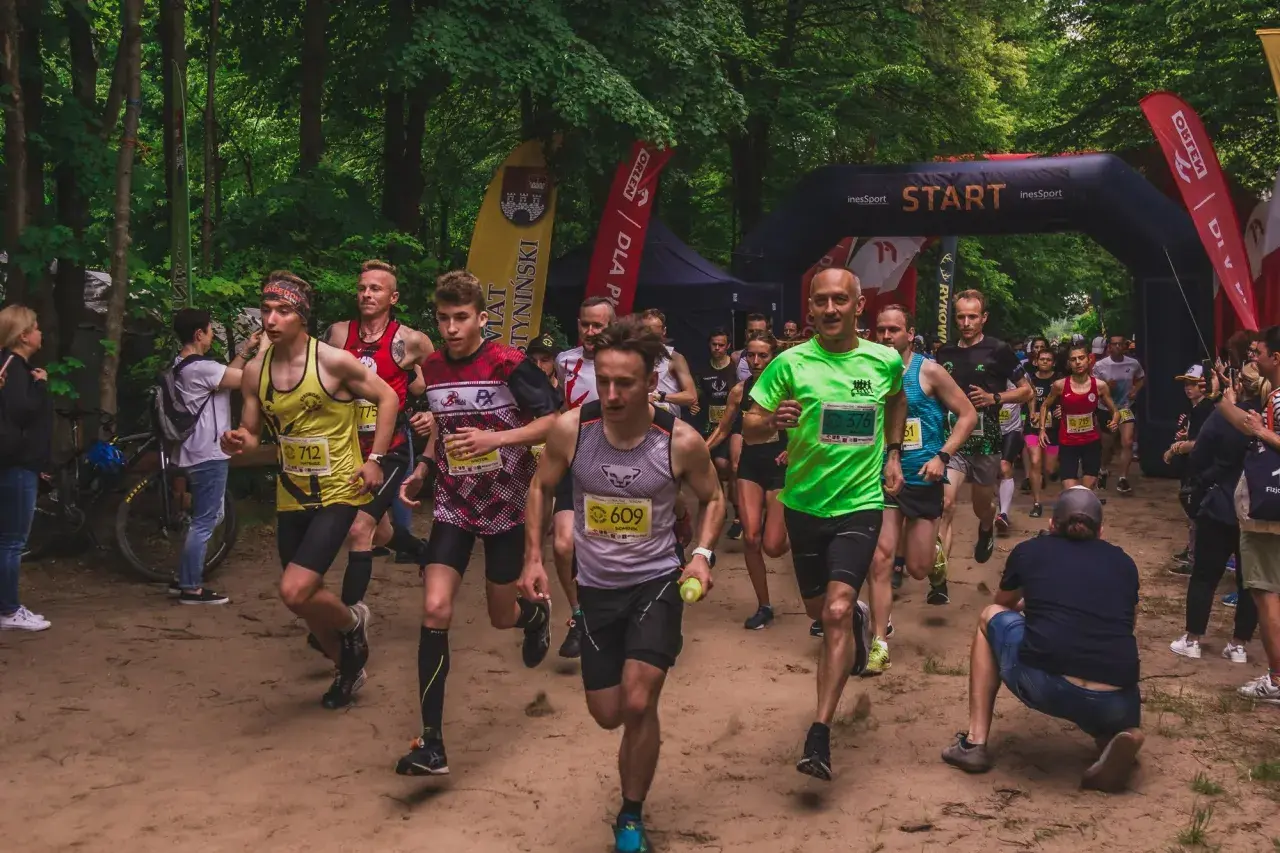 Uczestnicy biegu ultra trail startują w lesie. Widać łuk startowy, flagi i biegaczy w kolorowych koszulkach.