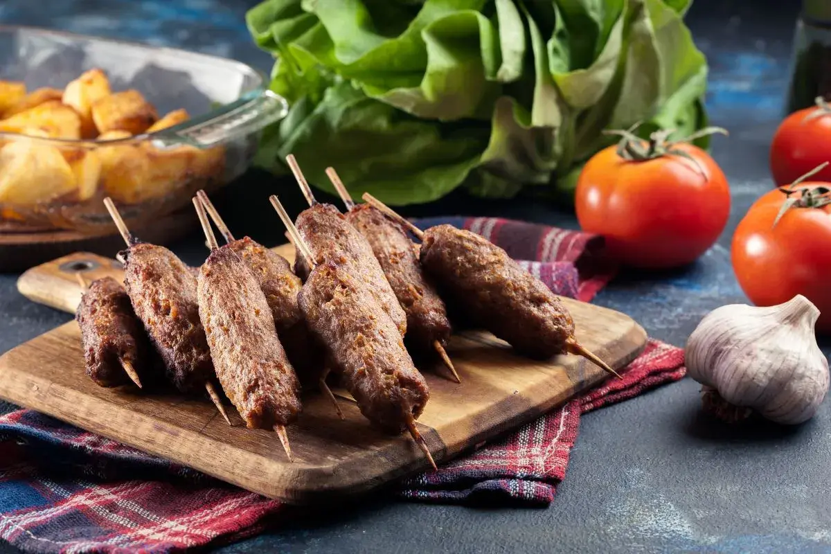 Aksamitne ćevapi na drewnianej desce, obok pomidory, czosnek i pieczone ziemniaki.