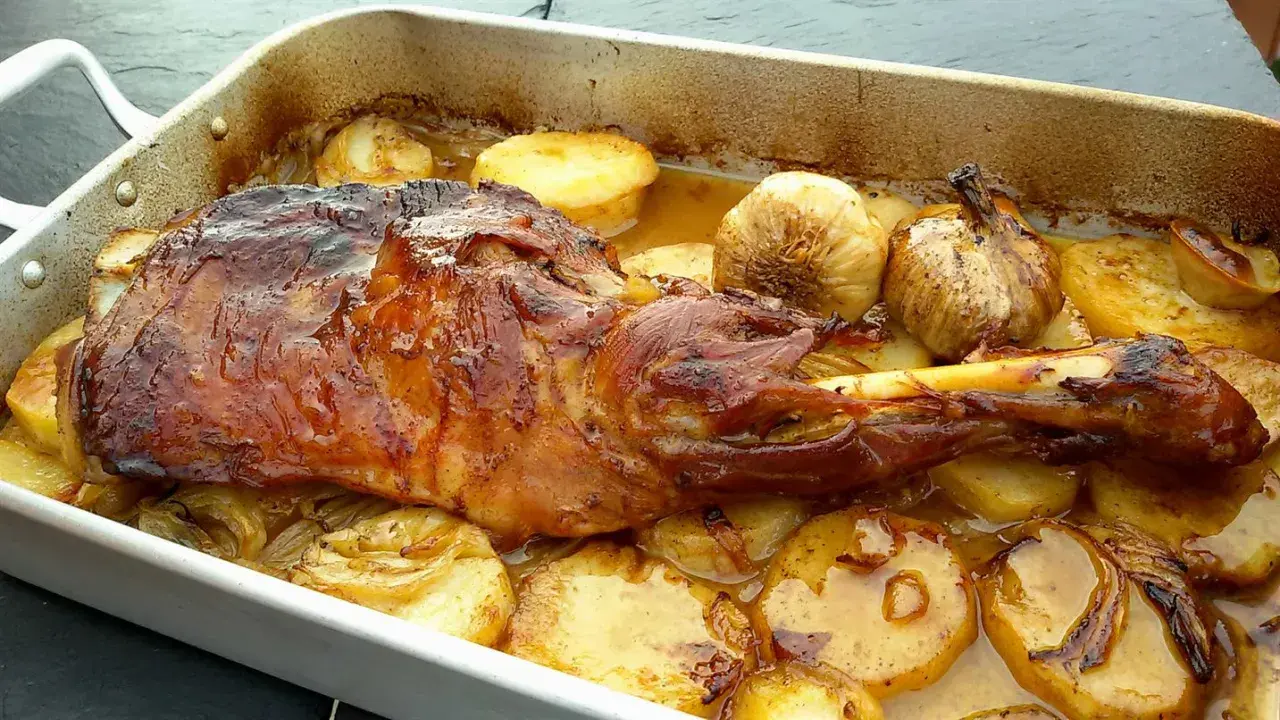 Paletilla de cordero al horno dorada y jugosa, acompañada de patatas asadas y ajos tiernos en una bandeja.
