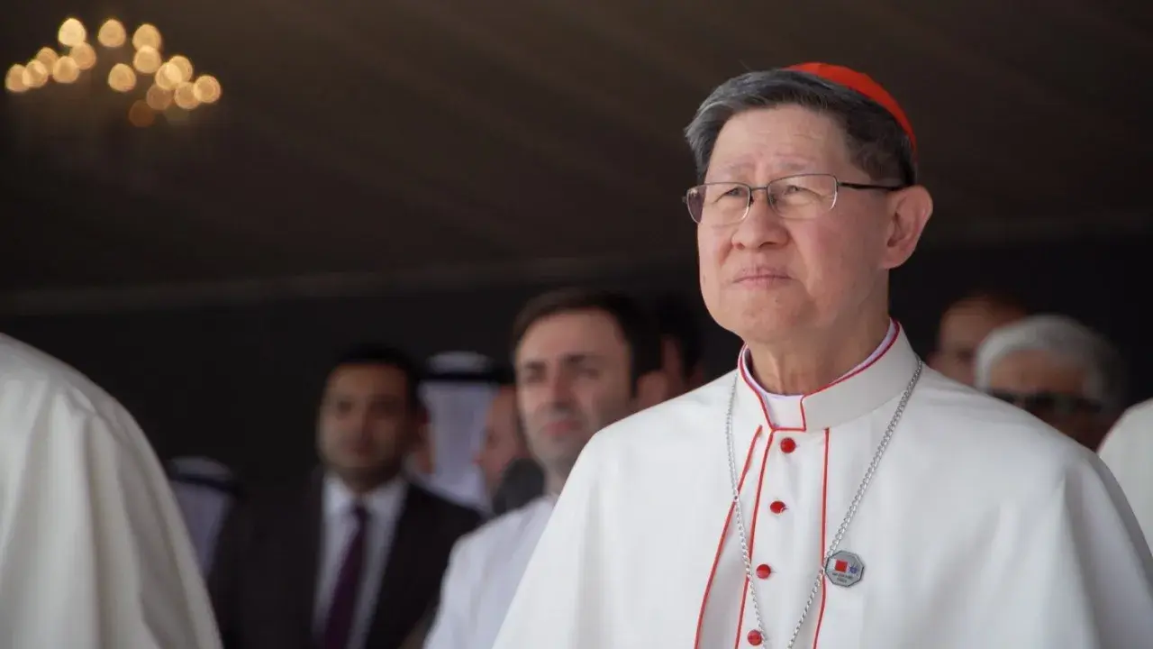 Kardinal Tagle, mit Brille und roter Pileolus, trägt ein weißes Gewand mit roten Akzenten. Im Hintergrund unscharfe Männer.