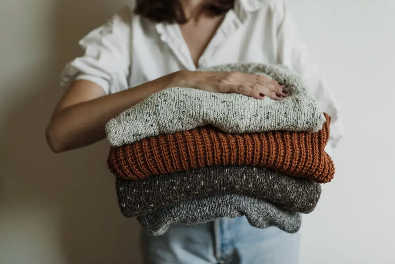 Eine Person hält einen Stapel gestrickter Pullover. Die Pullover sind in Grau-, Braun- und Beigetönen gehalten. Sie sind bereit, um **Wolle bei 40 Grad waschen** zu können.