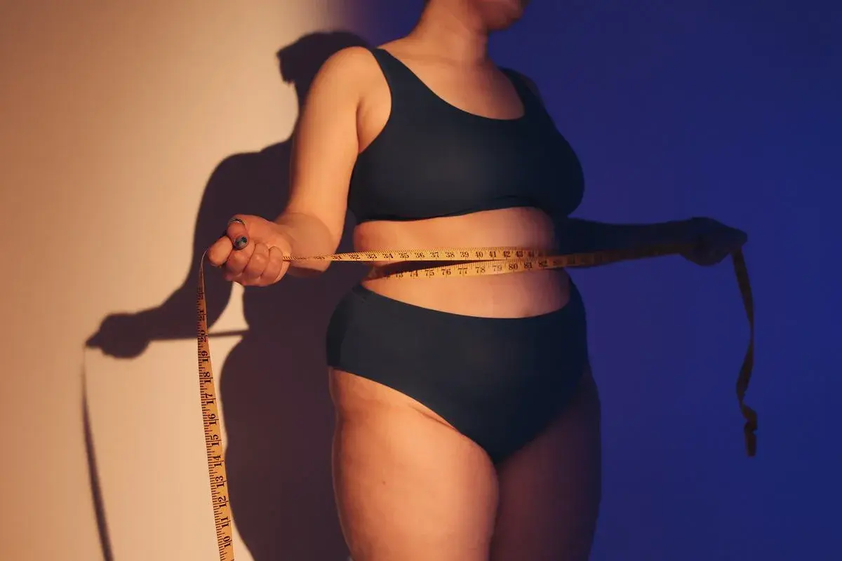 Osoba mierzy talię centymetrem. Ciemne światło podkreśla kształty, wywołując dyskomfort związany z body shaming.