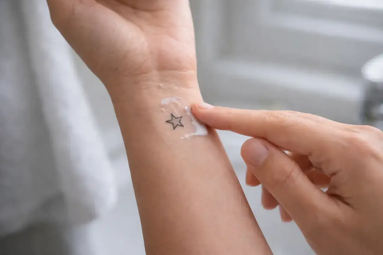 Un doigt applique une crème sur un petit tatouage en étoile sur le poignet. Le tatouage piqué trop profond semble légèrement irrité sous la crème.