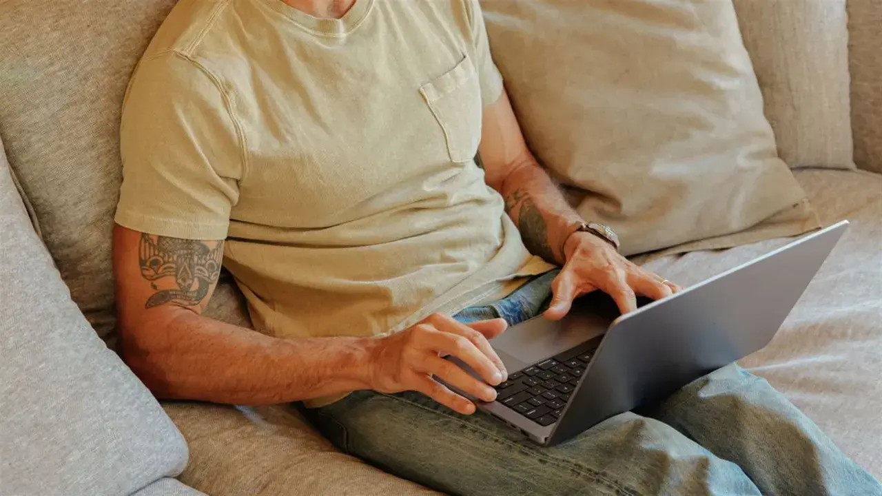 Man med tatuering på armen sitter i soffan med laptop, kanske för att han är hemma under en ny sjukperiod.