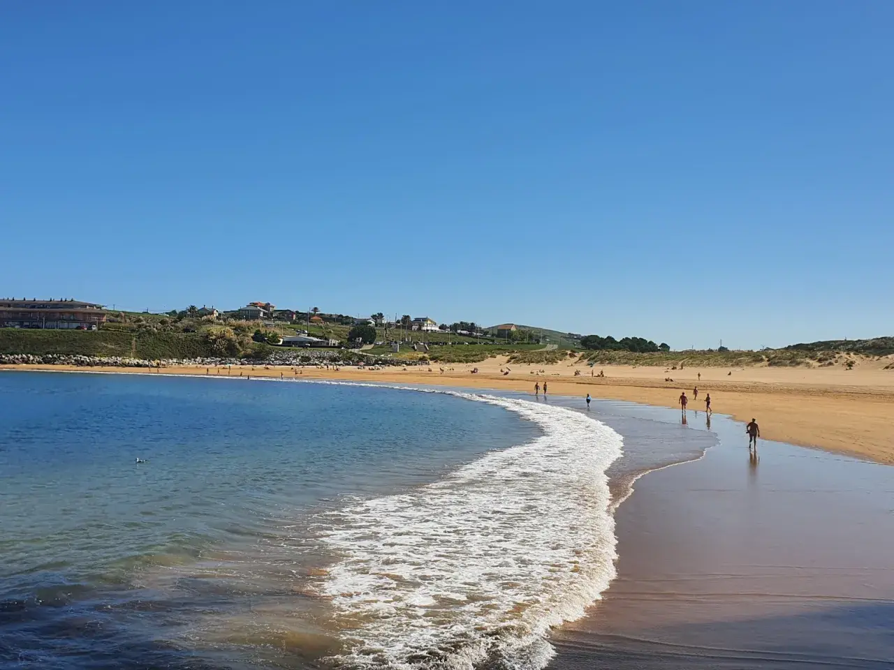 La **playa de Cuchía** se extiende bajo un cielo azul, con gente paseando por la arena dorada y el mar acariciando la costa.