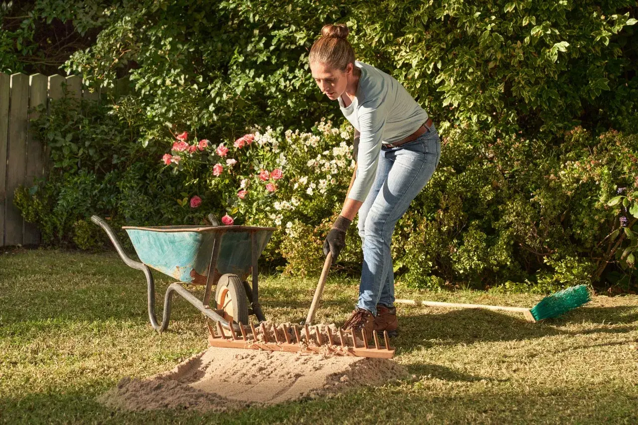 Frau verteilt mit Harke Sand auf dem Rasen. Eine Schubkarre und ein Besen liegen daneben.