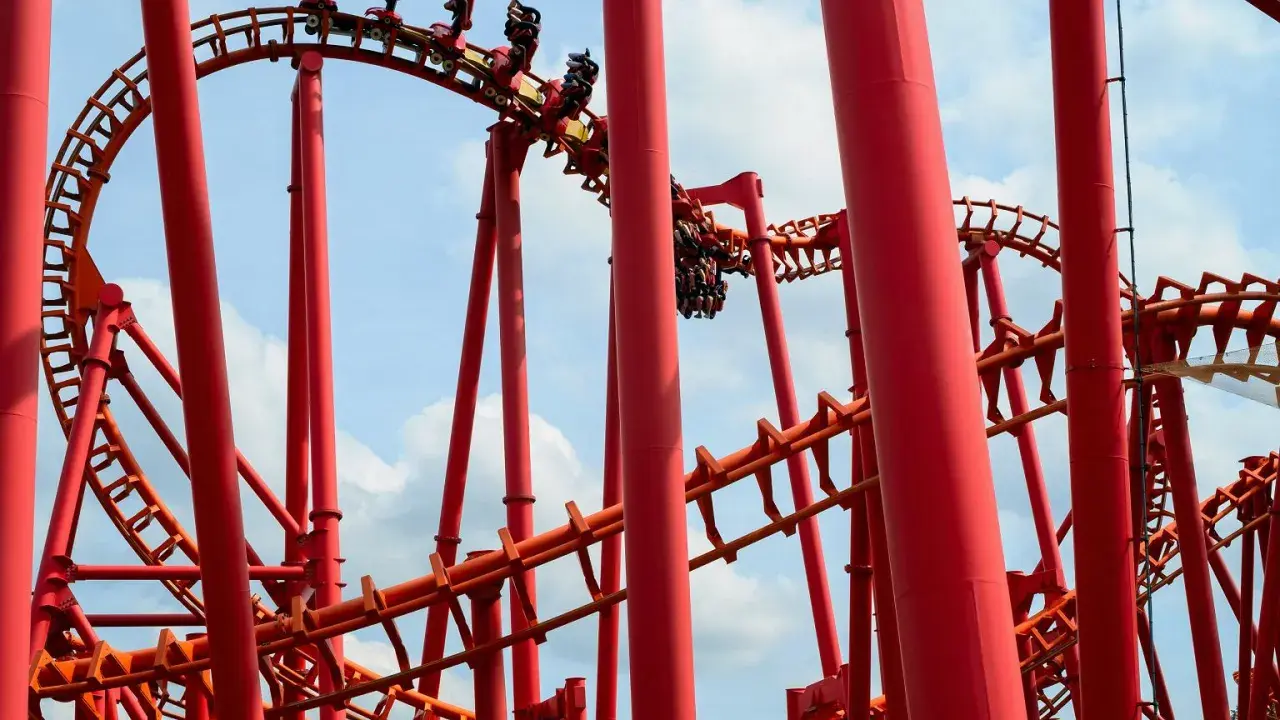 Czerwona kolejka górska z ludźmi w wagonikach na tle błękitnego nieba z chmurami.