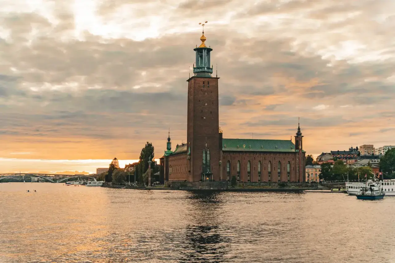 Stockholm Stadshus vid solnedgången, en vy som kunde vara från stockholm 1900. Vatten, båtar och en bro i bakgrunden.