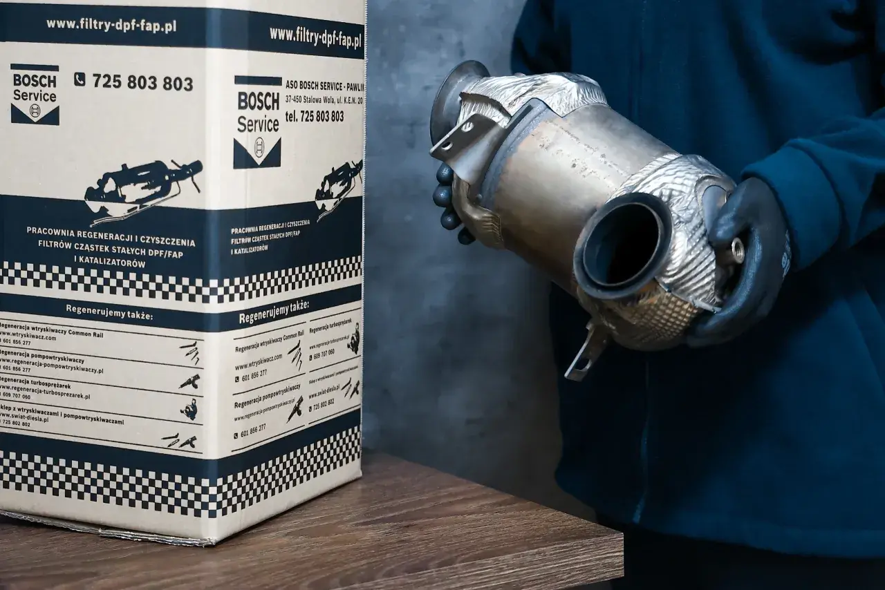 Osoba w rękawiczkach trzyma element układu wydechowego, prawdopodobnie filtr DPF. Obok stoi karton z logo Bosch Service, informujący o regeneracji filtrów DPF w dieslu.