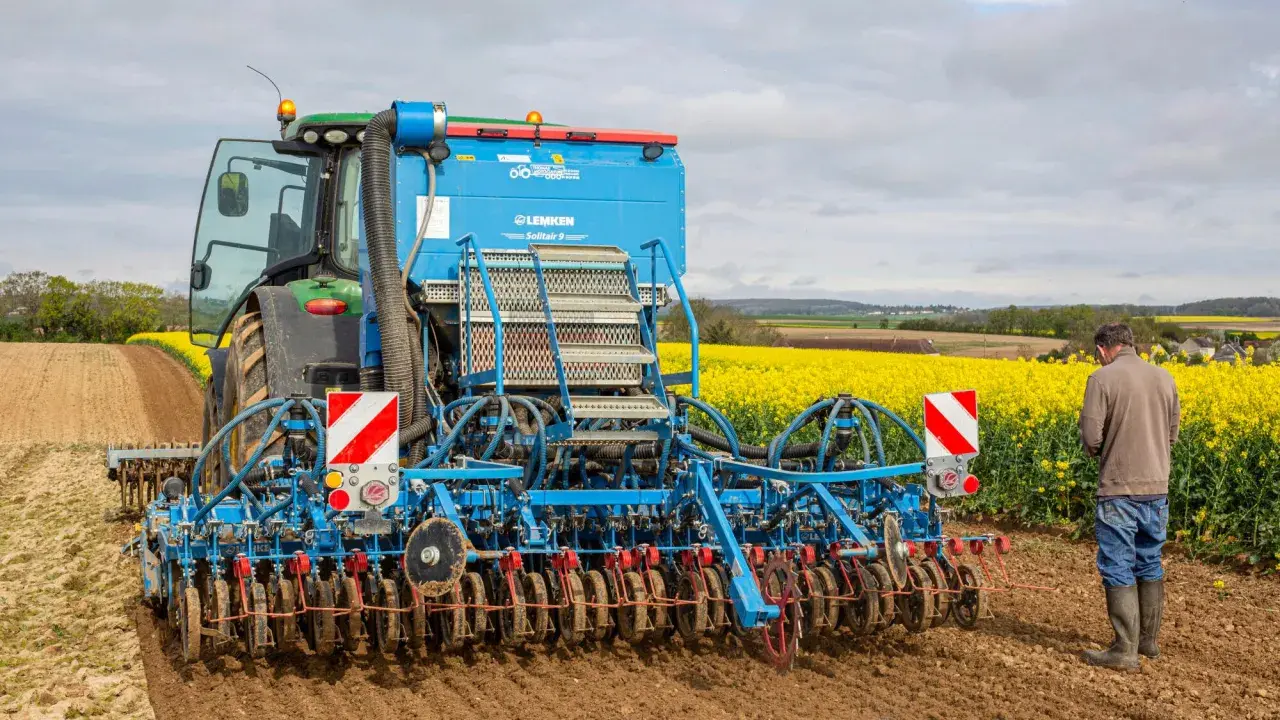 Un tracteur vert et une semoir Lemken Solitair 9 préparent le terrain pour le semis d'avoine de printemps, avec un champ de colza en fleurs en arrière-plan.