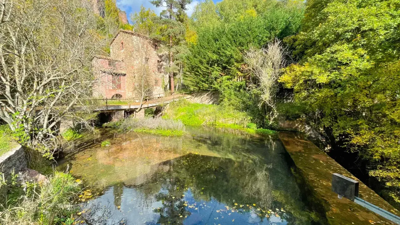 Antigua edificación de piedra junto a un río cristalino, rodeada de frondosa vegetación otoñal. Un puente de madera cruza el agua, conectando con las **fonts del Llobregat**.