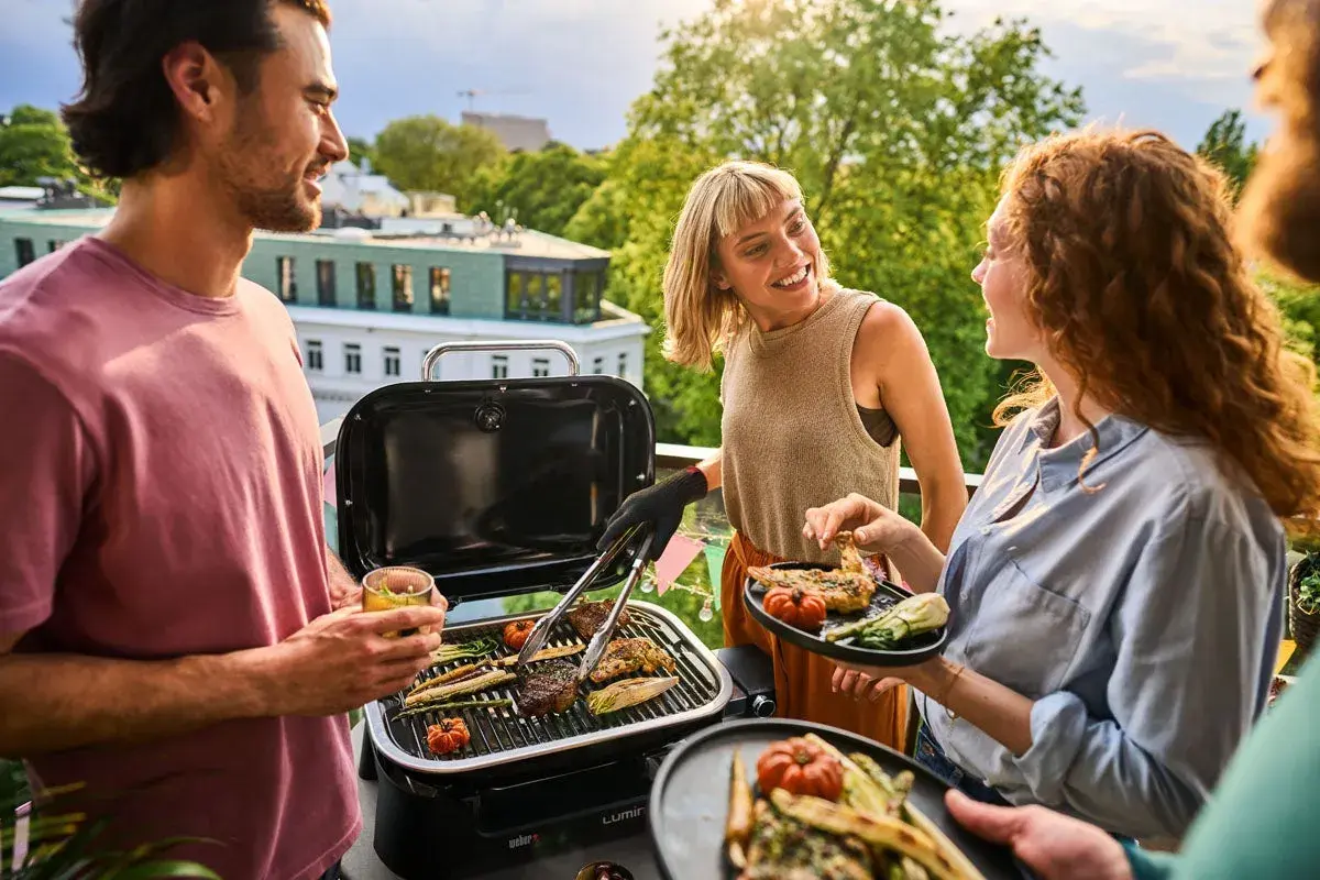 Vänner njuter av en grillfest på balkongen med en bärbar kolgrill. Saftiga biffar och grönsaker grillas medan solen går ner.