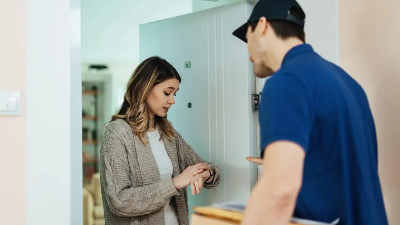 Eine Frau prüft ihre Uhr, während ein Lieferbote mit einem Paket vor der Tür steht. Die Zahlung avisiert, die Lieferung ist bald da.