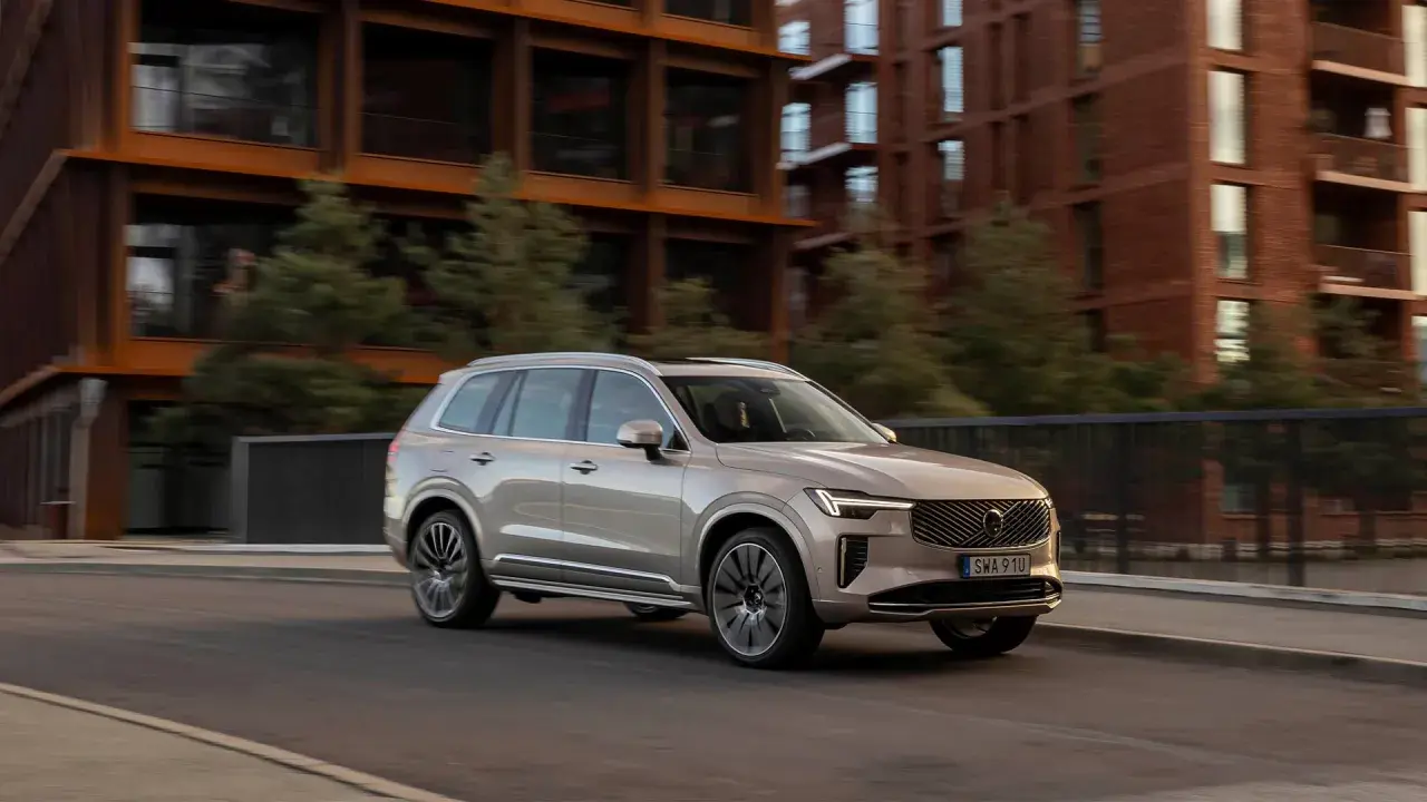 En ljusbeige Volvo SUV kör på en stadsgata. Bakgrunden visar moderna tegelbyggnader och gröna träd. Bilen visar upp olika volvo utrustningsnivåer.