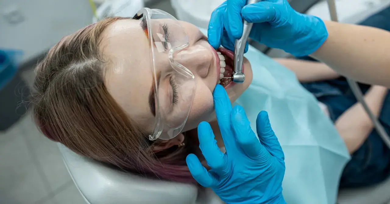 Dentysta w niebieskich rękawiczkach wykonuje zabieg, który wyjaśnia, co to jest leczenie kanałowe. Pacjentka ma okulary ochronne.