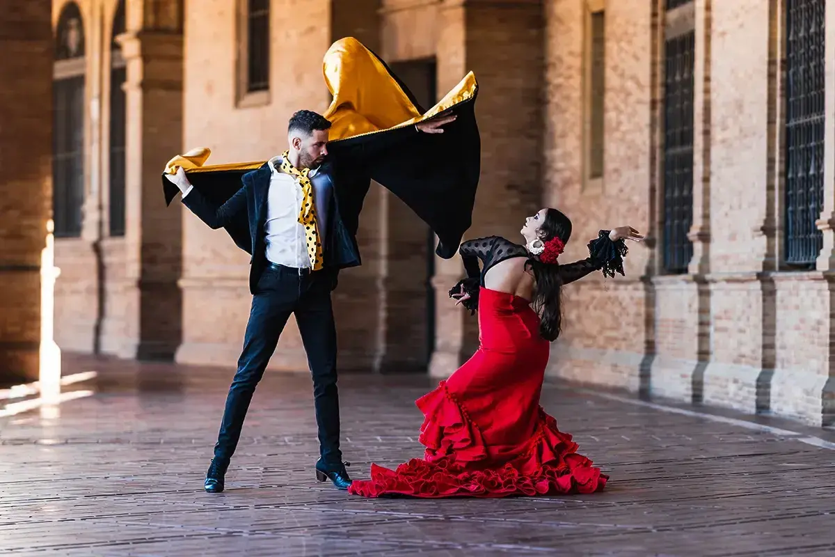 Pareja bailando flamenco con pasión. Él, con capa negra y dorada, ella, con vestido rojo voluminoso. Puro jaleo flamenco.