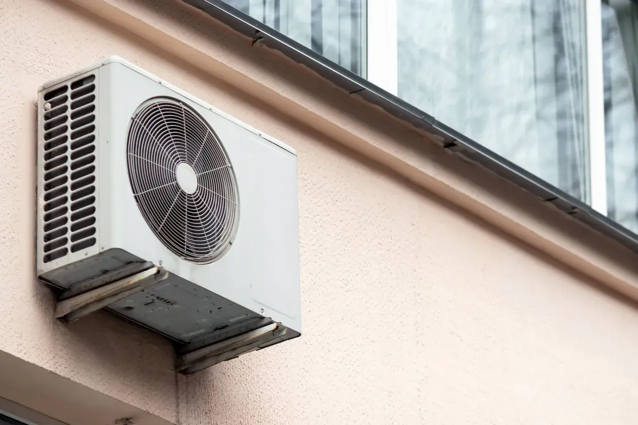 Klimatyzacja w bloku, biała jednostka zewnętrzna zamontowana na jasnoróżowej ścianie, z widocznym wentylatorem i kratką.