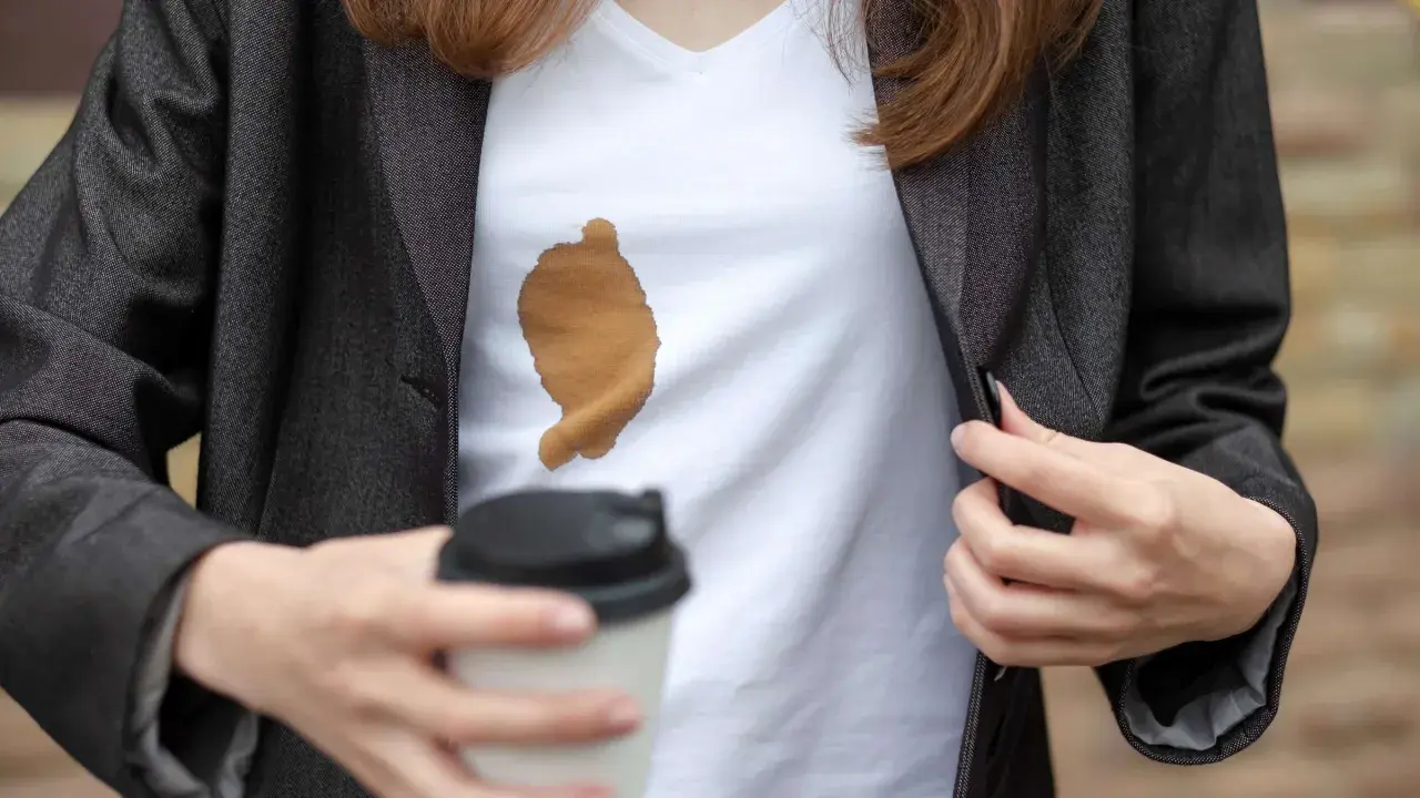 Frau mit Kaffeefleck auf weißem T-Shirt unter dunklem Blazer. Ein Becher Kaffee in der Hand. Tipps, wie man kaffeeflecken aus kleidung entfernen kann.