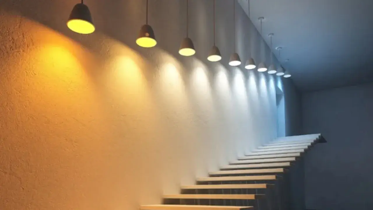 Escalera con luces colgantes que crean un degradado de luz y color, desde cálido hasta frío, iluminando la pared texturizada.