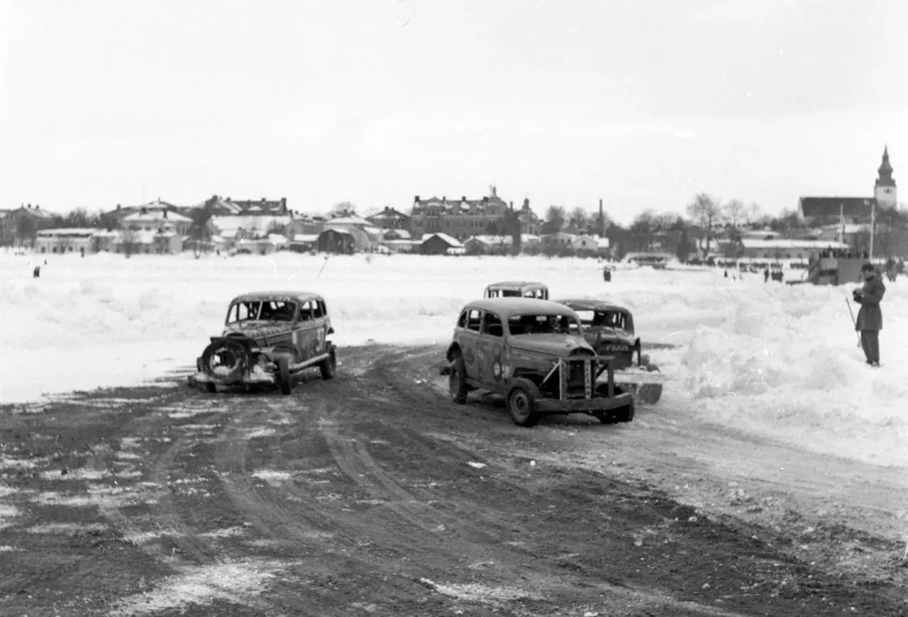 Snöig vinterdag med flera 70 tals bilar redo för race. En åskådare står vid sidan av banan.