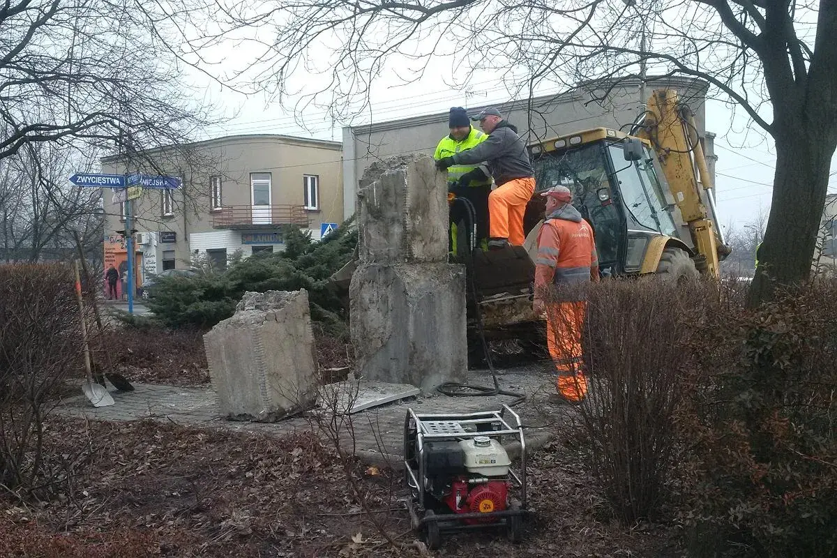 Prace przy pomniku, gdzie widać objawy zapadania się ziemi. Pracownicy w pomarańczowych strojach i koparka próbują wzmocnić podłoże.