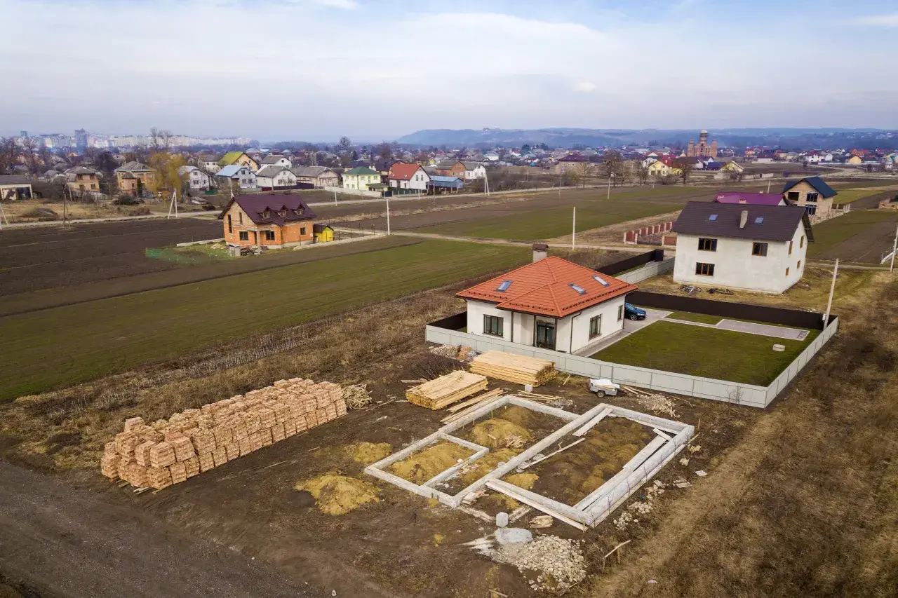 Działka inwestycyjna a budowlana: widać fundamenty domu, sterty cegieł i drewna, gotowe domy i pola uprawne w tle.