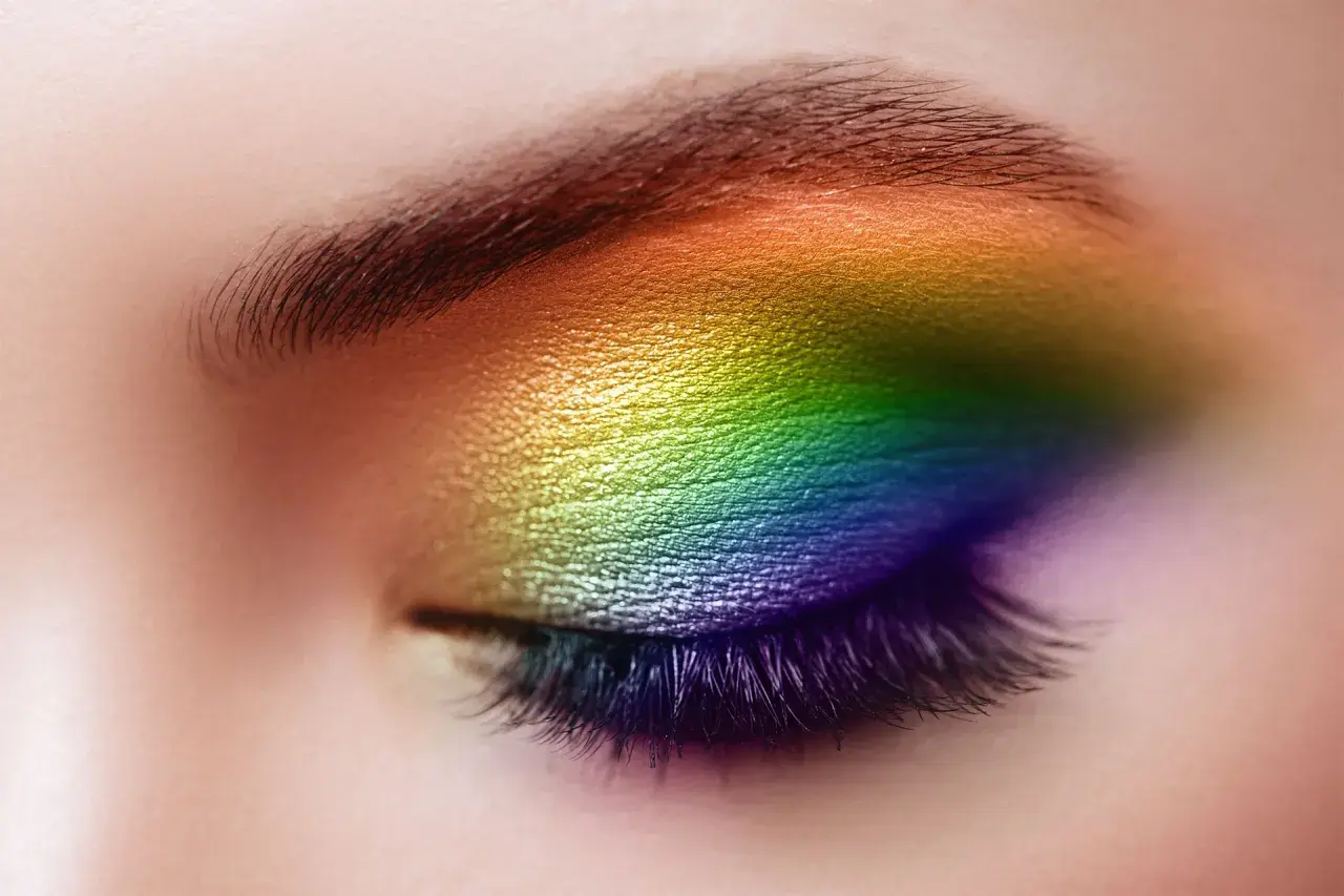Un œil fermé orné d'un maquillage arc-en-ciel vibrant, du jaune au violet, sublimant le regard. Un maquillage yeux bleus audacieux et coloré.