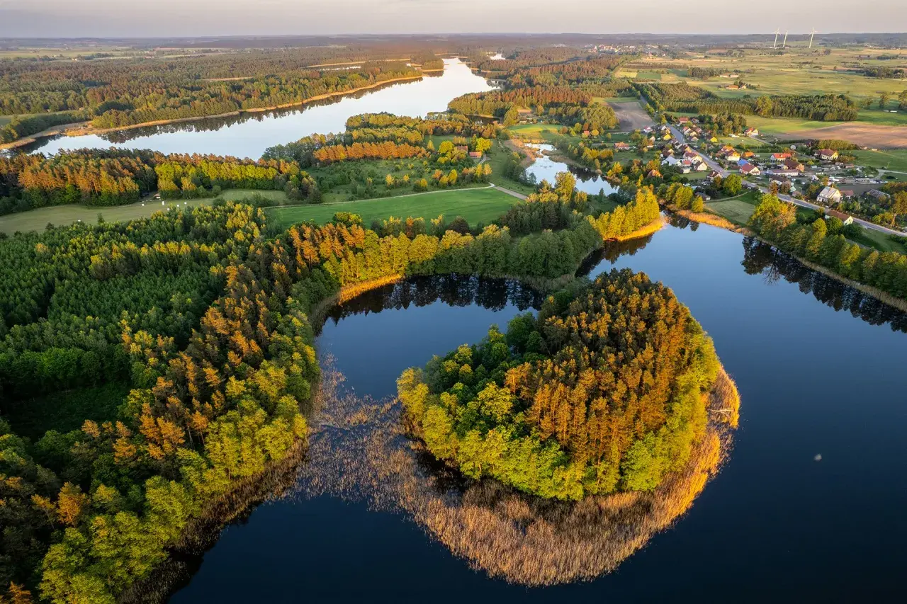 Widok z lotu ptaka na jezioro otoczone lasem i małą wioskę na Mazurach.