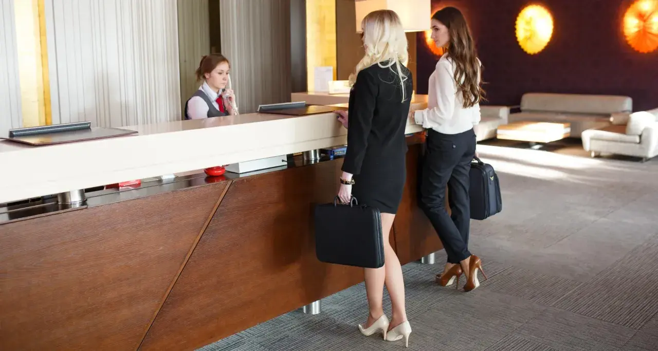 La receptionist accoglie i clienti che arrivano in albergo. Due donne d'affari con valigette attendono al bancone.