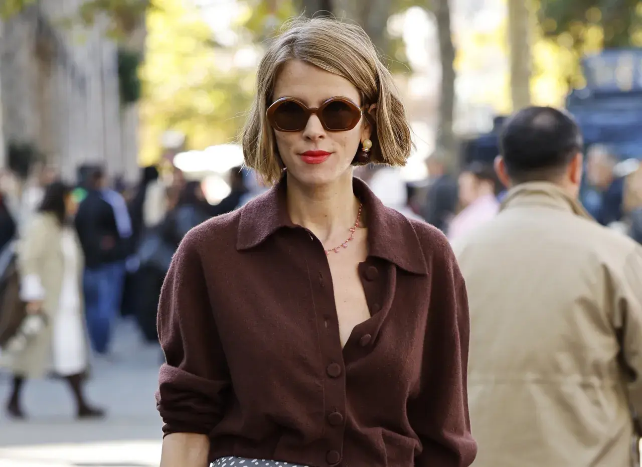 Mujer con corte francés con raya lateral, gafas oscuras y labial rojo, luciendo un suéter marrón.