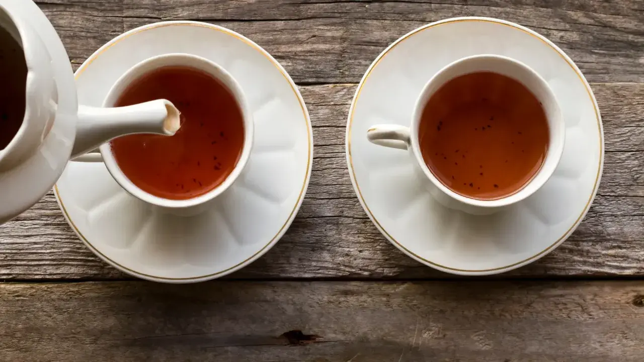 Dwie filiżanki z herbatą pu erh, z których jedna jest nalewana z czajniczka. Napój ma głęboki, bursztynowy kolor, a jego właściwości zdrowotne są cenione od wieków.