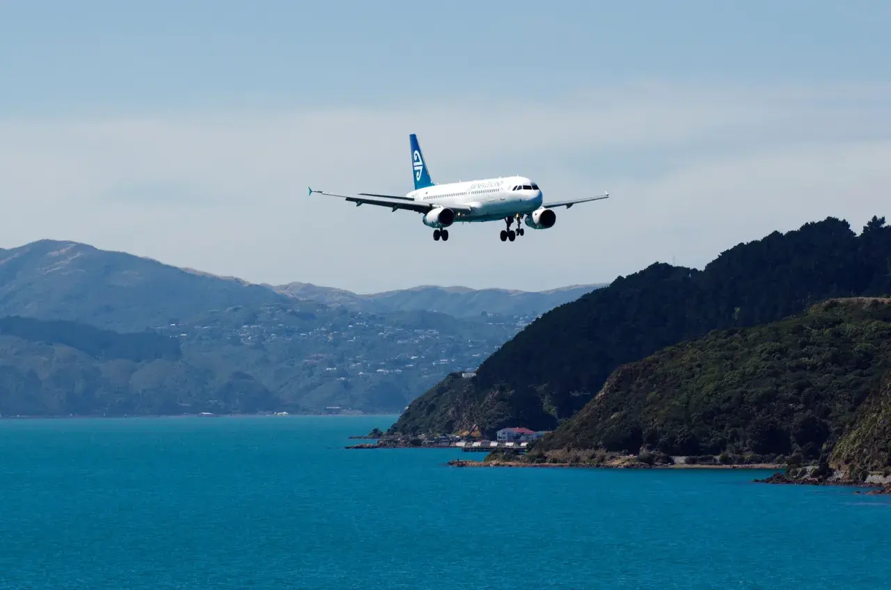 Samolot Air New Zealand ląduje nad turkusową wodą, zbliżając się do wybrzeża. Zastanawiasz się, ile kosztuje lot do Nowej Zelandii?