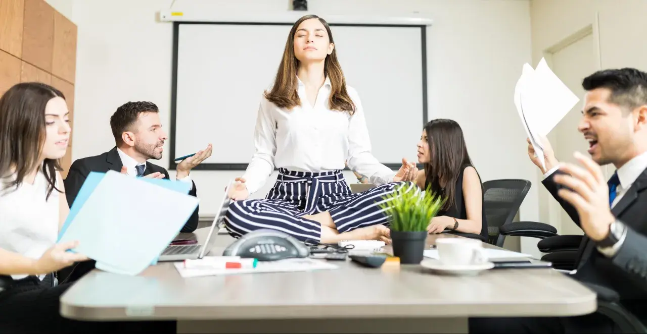 A woman meditates in lotus pose at a conference table while colleagues argue heatedly around her, creating a stark contrast between chaos and calm conversation.