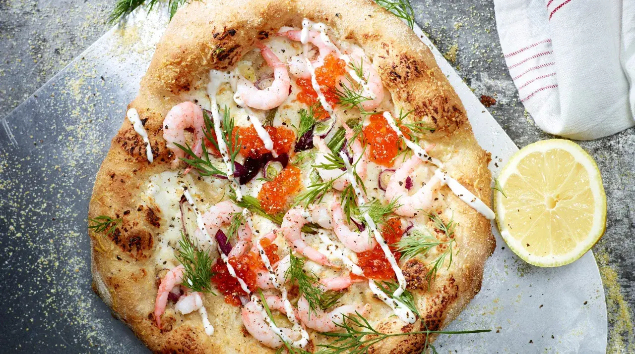 En läcker pizza med räkor och mozzarella, toppad med rom och dill. En citronskiva ligger bredvid.