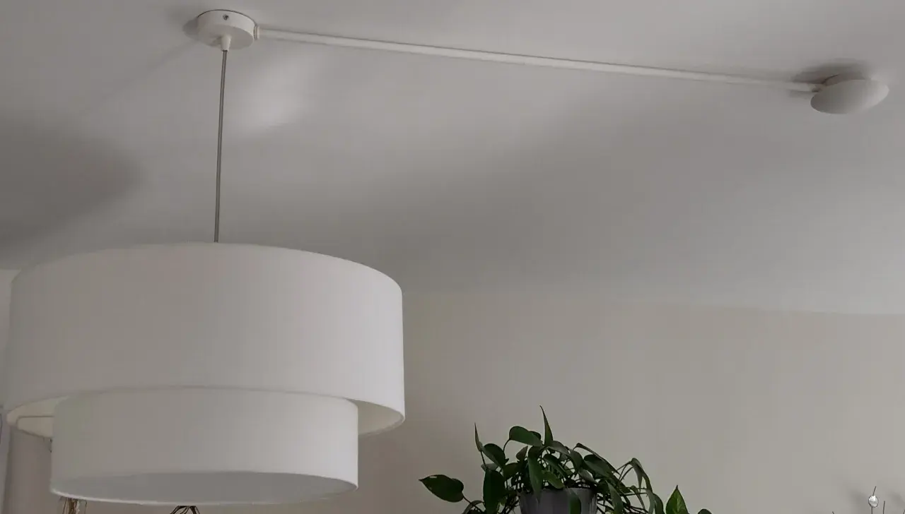 Lámpara de techo con un punto de luz desplazado, colgada de un riel blanco. Una planta verde asoma por la derecha.