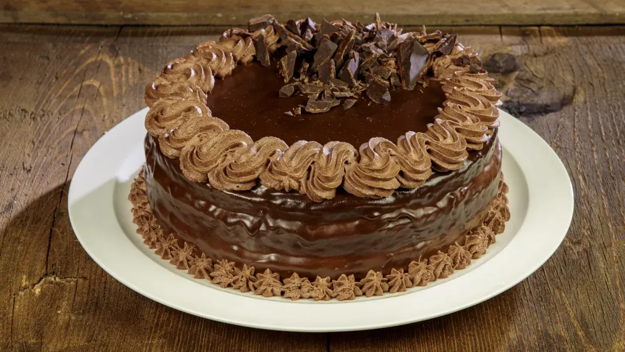 Torta compleanno cioccolato e panna, ricoperta di glassa lucida e decorata con riccioli di panna e scaglie di cioccolato.