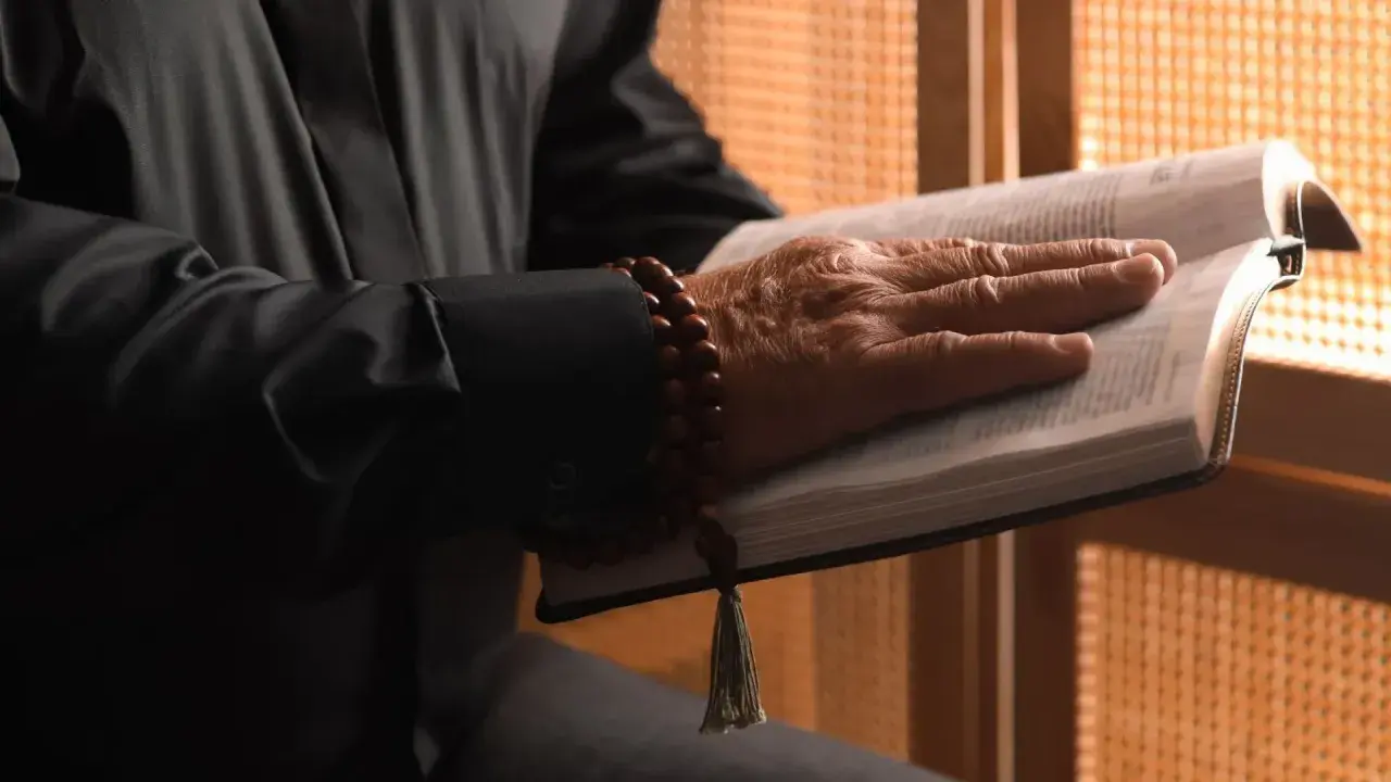 Dłoń starszego mężczyzny spoczywa na otwartej Biblii. Wokół nadgarstka ma drewniane paciorki. Gdzie jest pastor?