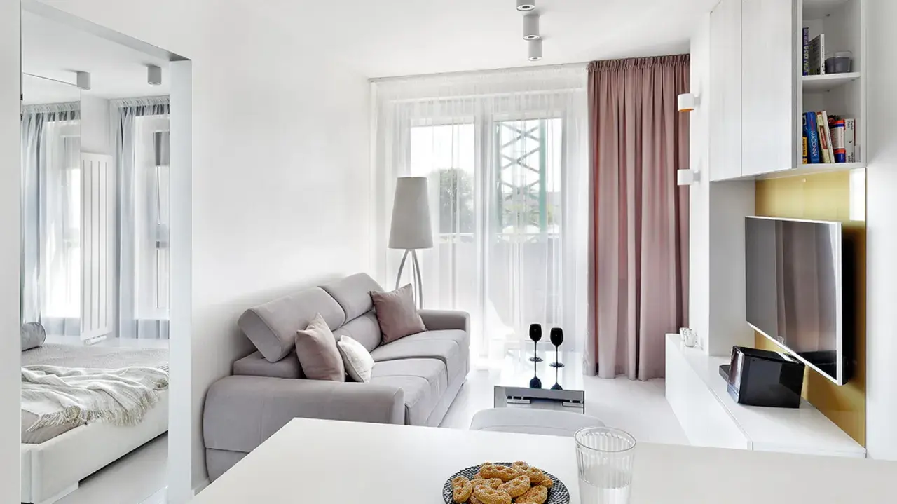 Jasny, bardzo mały salon w bloku z szarą kanapą, stolikiem kawowym i widokiem na balkon. Obok widać łóżko.