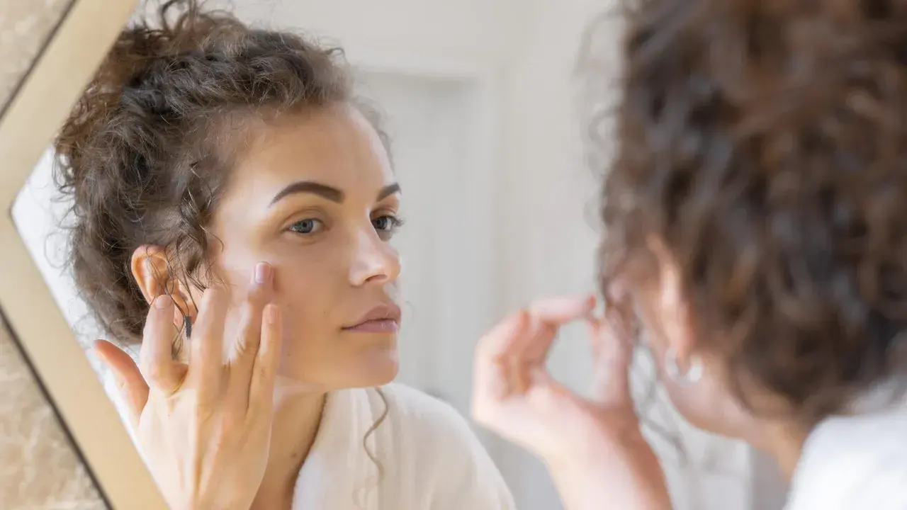 Woman applying cream to her face in a mirror.