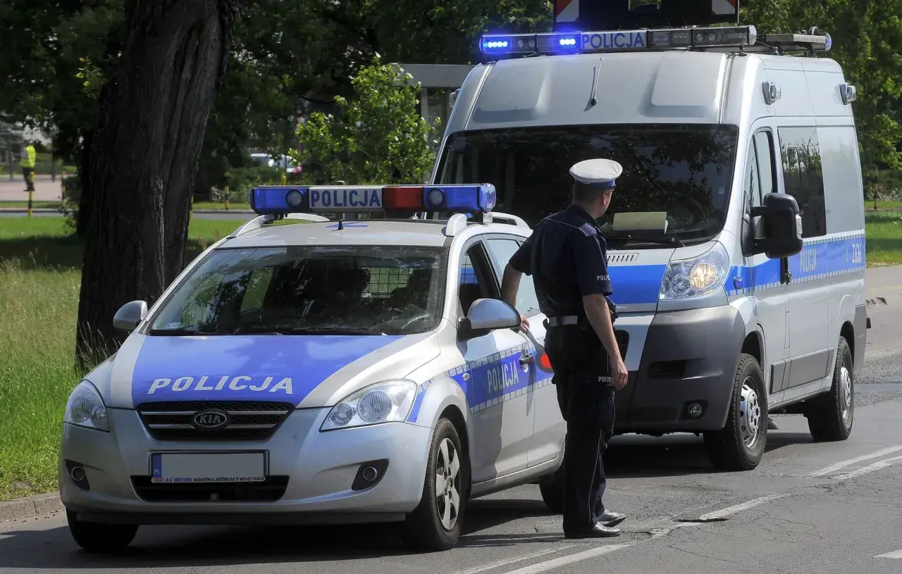 Policjant stoi obok radiowozu i furgonetki policyjnej z włączonymi sygnałami świetlnymi.