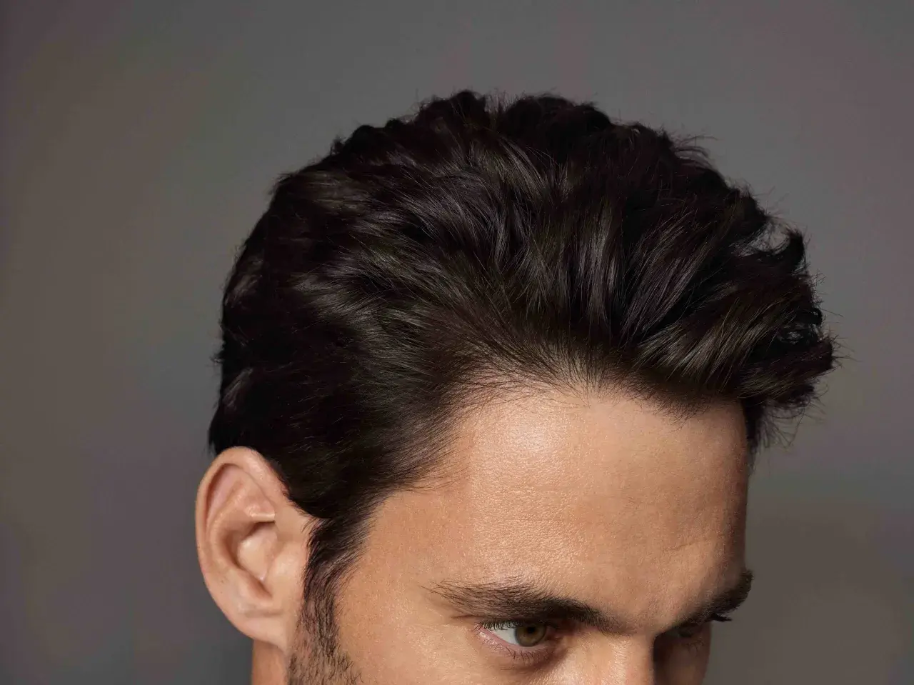 Primer plano de un hombre con mas cantidad de pelo oscuro y peinado hacia atrás, mostrando su oreja y parte de su rostro.
