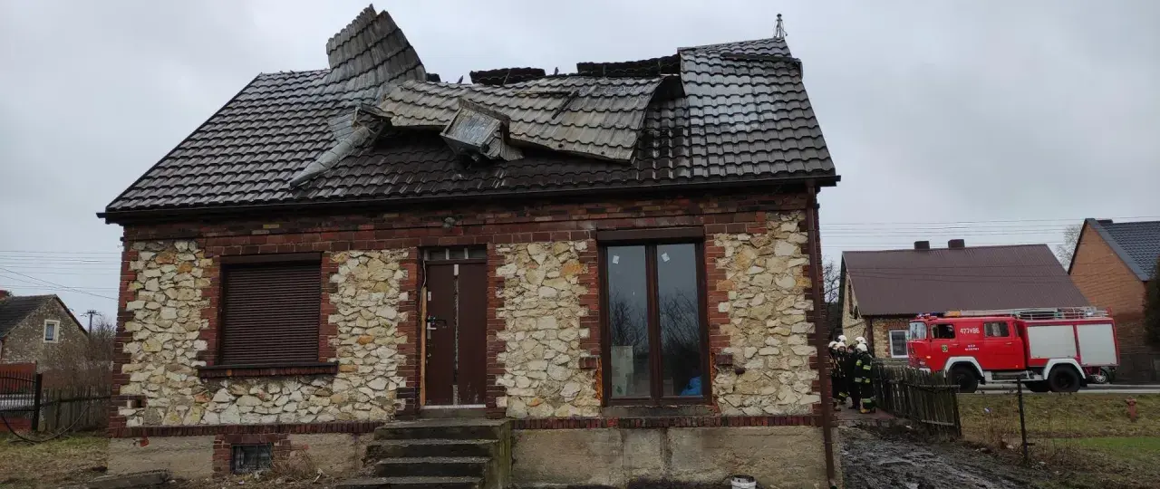 Zniszczony dach domu z kamienia wapiennego. Obok stoi wóz strażacki, a przy nim strażacy.