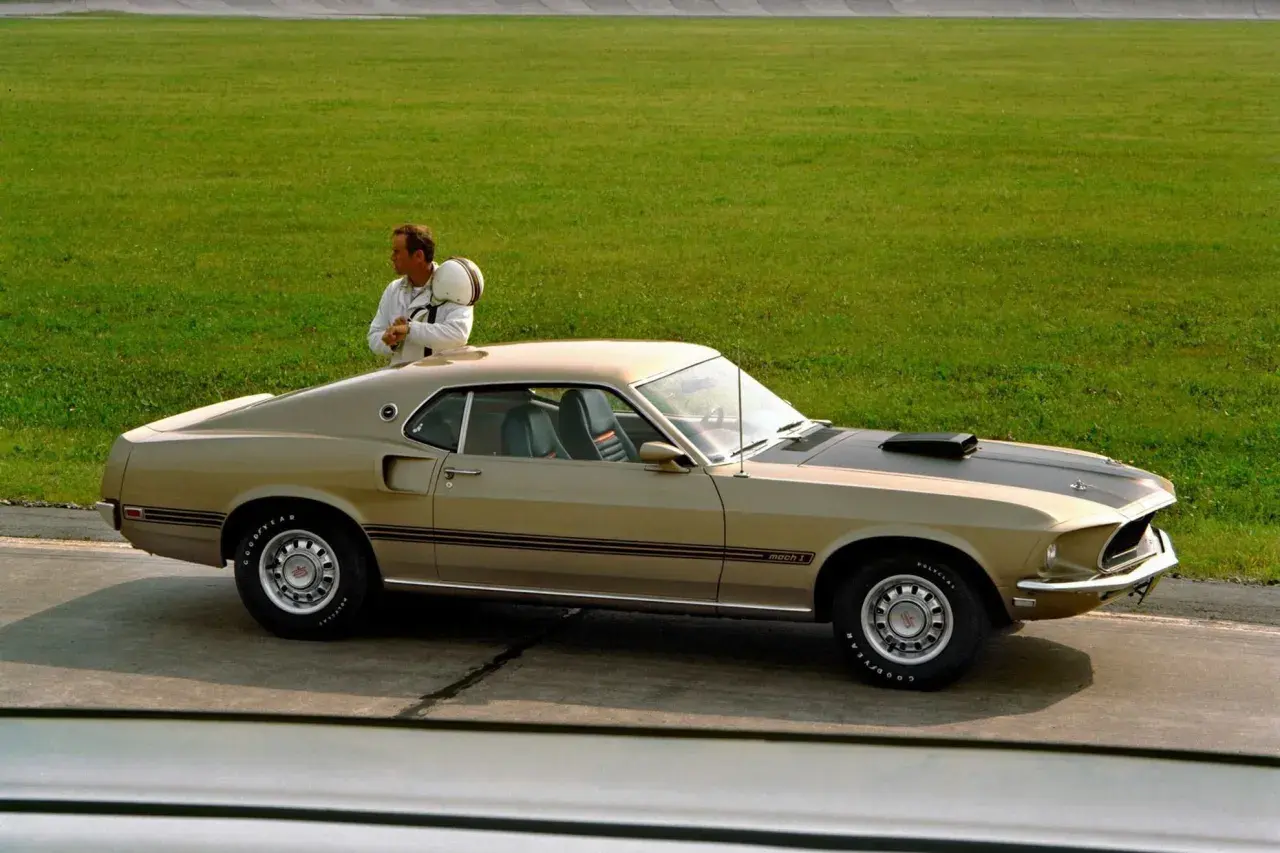 En man med hjälm lutar sig mot en guldig mustang 60 tal på en bana.