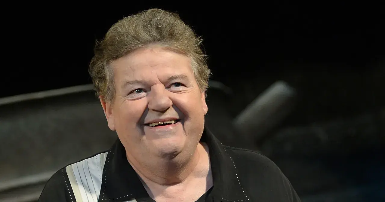 Uśmiechnięty Robbie Coltrane, który grał Hagrida, w czarnej koszuli z białymi pasami.