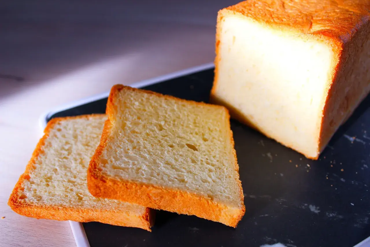 Pain de mie moelleux, parfait pour une recette pain de mie boulangerie maison. Deux tranches dorées et un pain entier sur un plateau sombre.